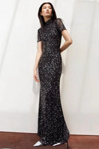 Woman in a black sequined gown with a high neckline and short sleeves, posing against a white wall.