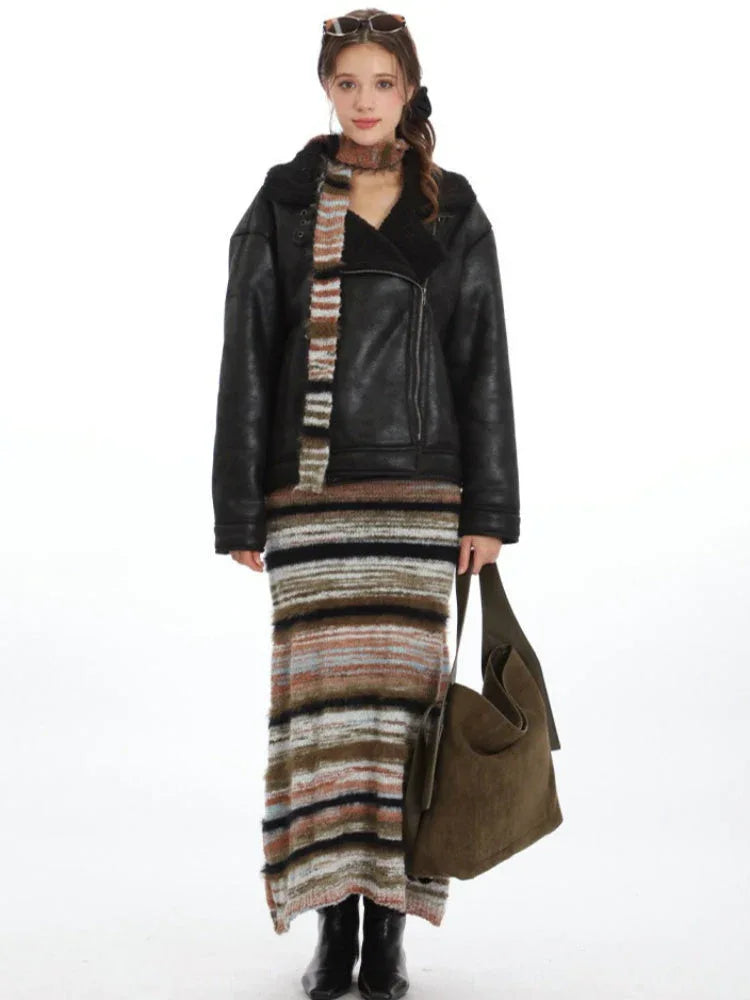 Model wearing a black faux leather jacket, a striped knit maxi skirt, and a multi-colored striped scarf, holding a green shoulder bag.