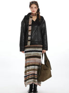 Model wearing a black faux leather jacket, a striped knit maxi skirt, and a multi-colored striped scarf, holding a green shoulder bag.