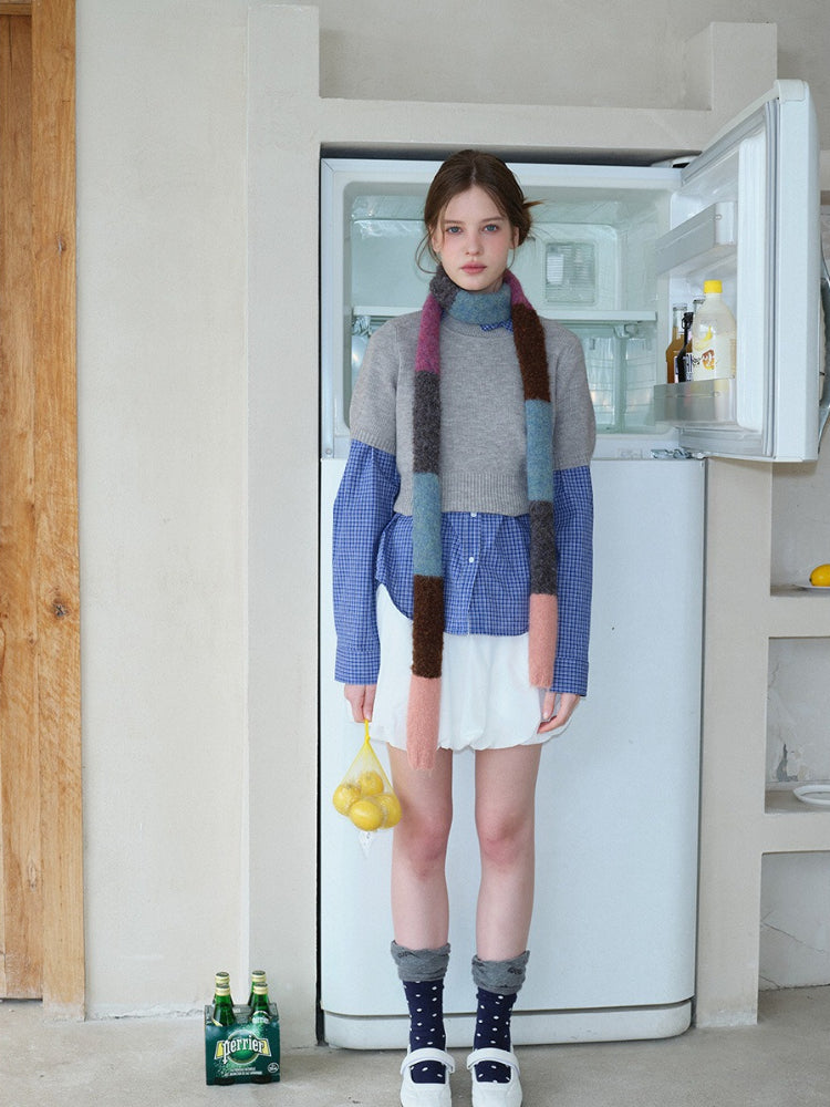 Model holding a mesh bag of lemons, wearing a grey cropped sweater over a blue plaid long-sleeved shirt, white bubble skirt, and a colorful striped scarf, in front of an open refrigerator.