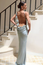 woman wearing a light green satin floor-length mermaid dress with a low back and floral embellishments, standing on a staircase