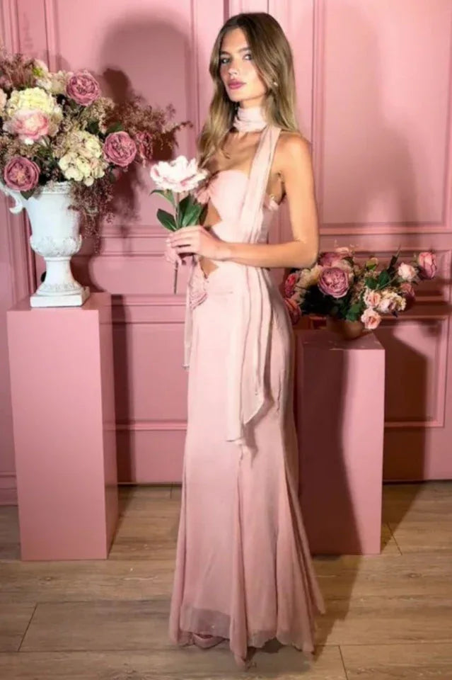 Woman wearing a pale pink halter neck maxi dress with cut-out details and a mermaid hemline, holding a flower and looking to the side in a pink studio setting.