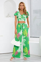 Woman wearing a green floral print two-piece set with a cropped V-neck top and wide-leg pants, holding a white handbag, front view.