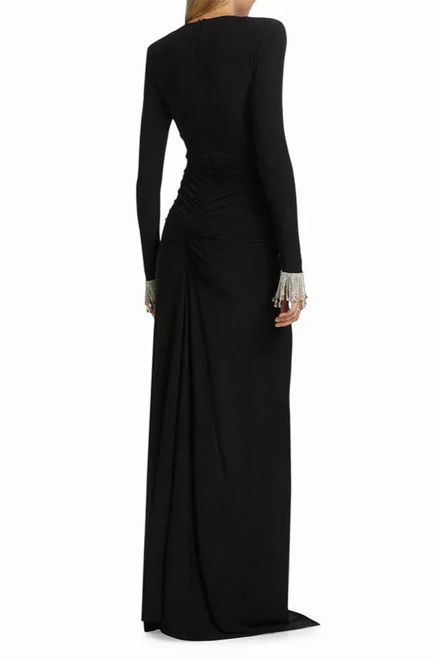 Back view of a woman wearing a long-sleeved black gown with a plunging V-neckline, ruched bodice, thigh-high slit, and embellished cuffs.