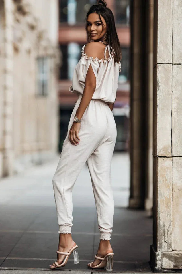 Woman from the back wearing a light-colored off-the-shoulder jumpsuit with a deep V-neck and tie shoulder straps, walking outdoors. Secondary image