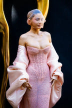 Close-up of a woman wearing a pink sequined strapless gown with oversized pink satin sleeves and a sheer head covering, posing against a dark background with a gold accent.