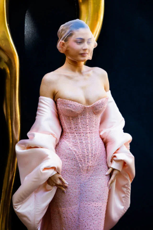Close-up of a woman wearing a pink sequined strapless gown with oversized pink satin sleeves and a sheer head covering, posing against a dark background with a gold accent.
