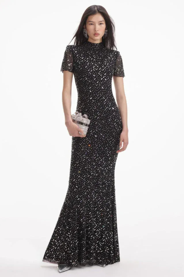 Model wearing a black sequined floor-length gown with a high neckline and short sleeves, with a silver clutch.