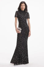 Model wearing a black sequined floor-length gown with a high neckline and short sleeves, with a silver clutch.
