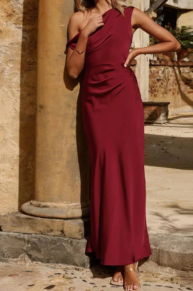 Woman posing in a burgundy one-shoulder satin maxi dress against a stone pillar.