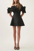Model wearing a black off-the-shoulder mini dress with large floral embellishments on the shoulders and black heels.
