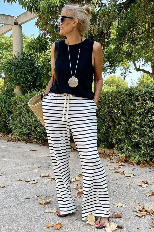 Woman from the side wearing a black sleeveless top and black and white striped wide leg pants, holding a straw tote bag.