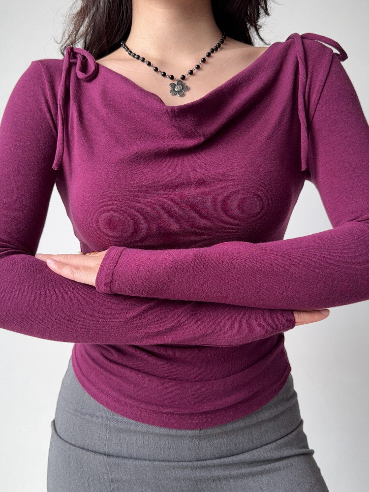 Model wearing a plum long-sleeve top with a draped neckline and ruched sides, arms crossed.