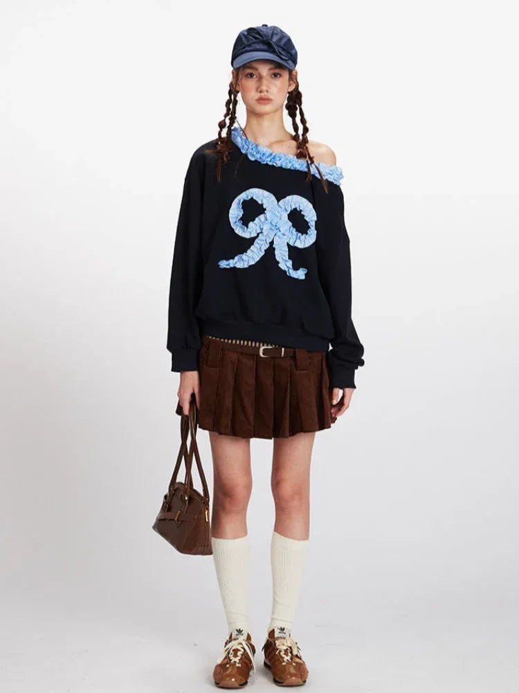 Model wearing a black off-the-shoulder sweatshirt with a large light blue appliqué bow on the front and ruffled blue trim.