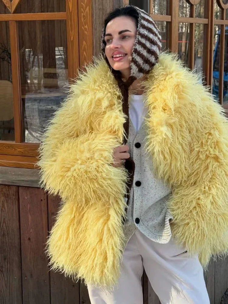 Woman wearing a yellow shaggy faux fur jacket over a white t-shirt and a grey cardigan with light-colored pants and a striped knit hat.