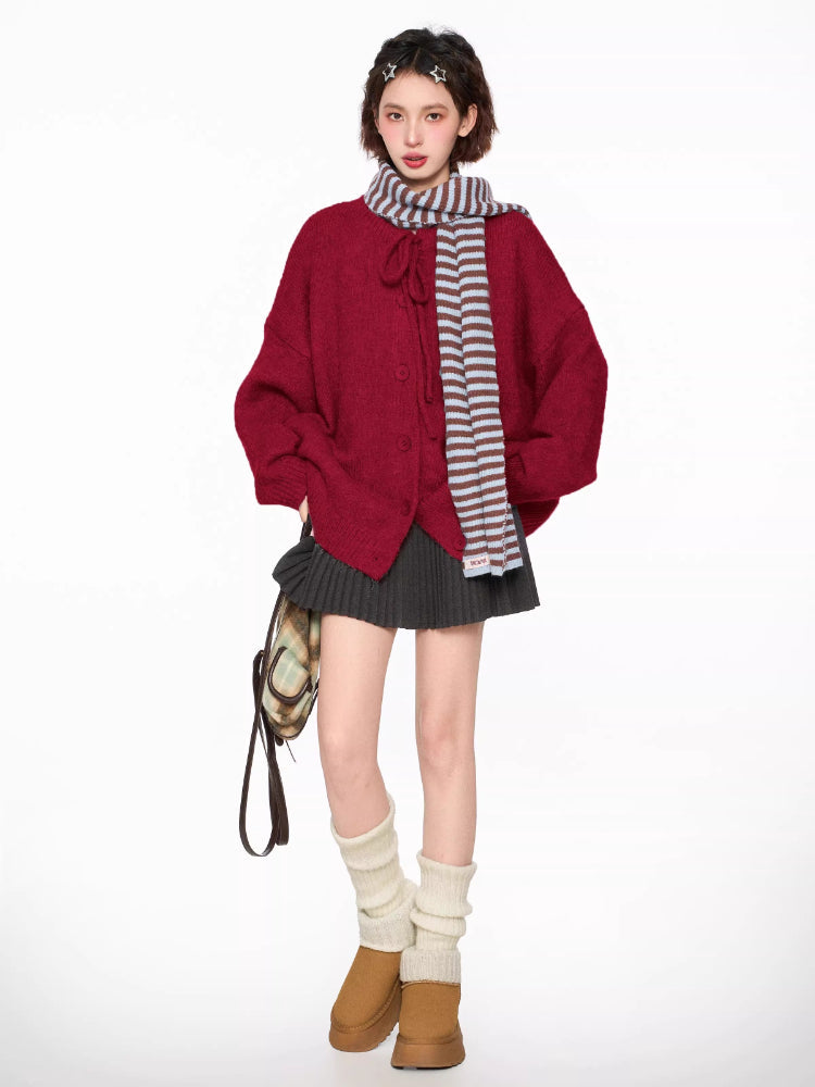 Woman wearing a red knit cardigan with tie neck and buttons, grey pleated skirt, brown crossbody bag, white leg warmers, and a striped scarf.