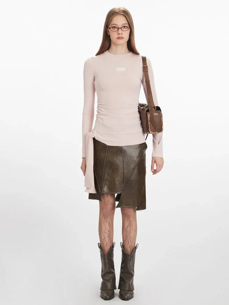 Full-body front view of a woman wearing a pink long-sleeve ruched top, a brown asymmetrical faux leather skirt, patterned tights, dark boots, and a brown shoulder bag.