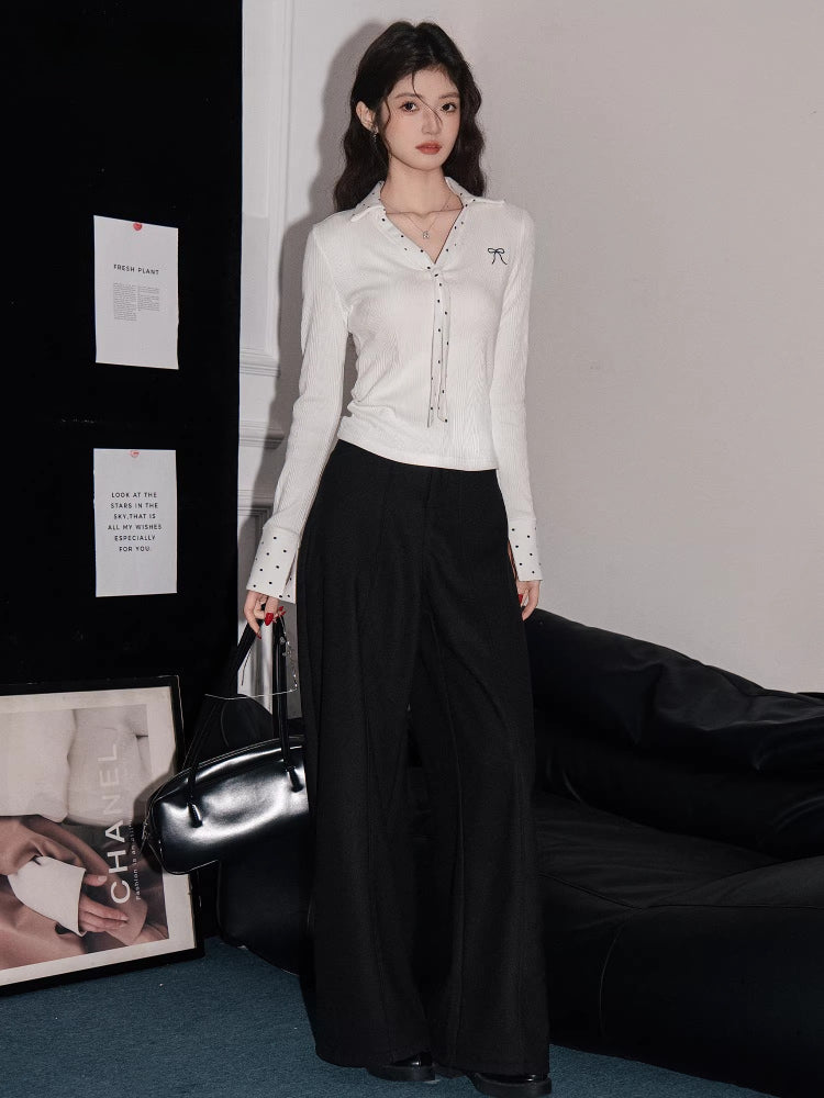 Full-body shot of a woman wearing a white ribbed long-sleeve shirt and black wide-leg pants, holding a handbag.