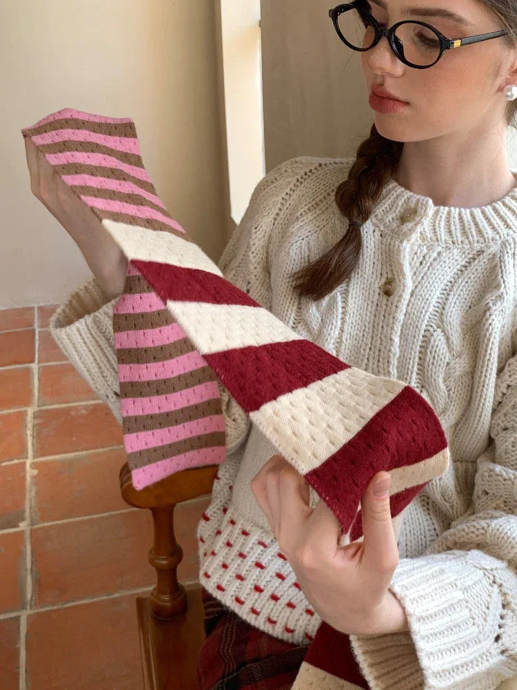 A young woman with glasses holding a striped scarf, wearing a cream cable knit cardigan.