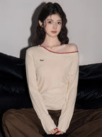 Woman wearing a cream off-the-shoulder long-sleeve top with red trim and brown pants, sitting on a black couch.