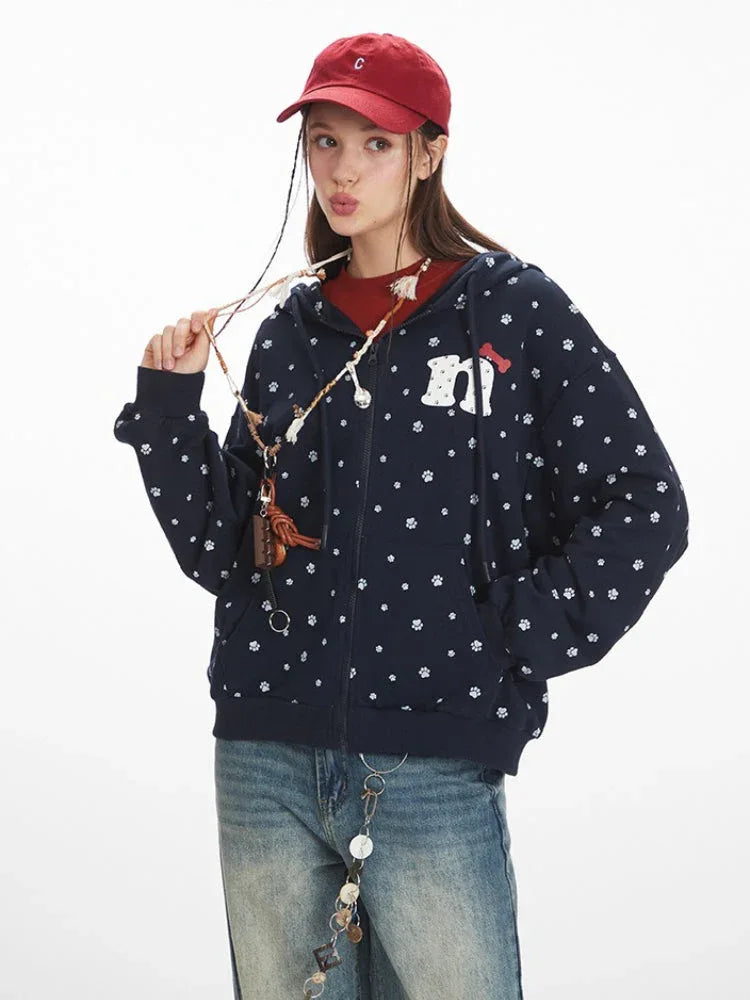 Front close-up of a woman wearing a navy blue hooded zip-up jacket with a white paw print pattern and a graphic detail on the chest, holding a necklace.