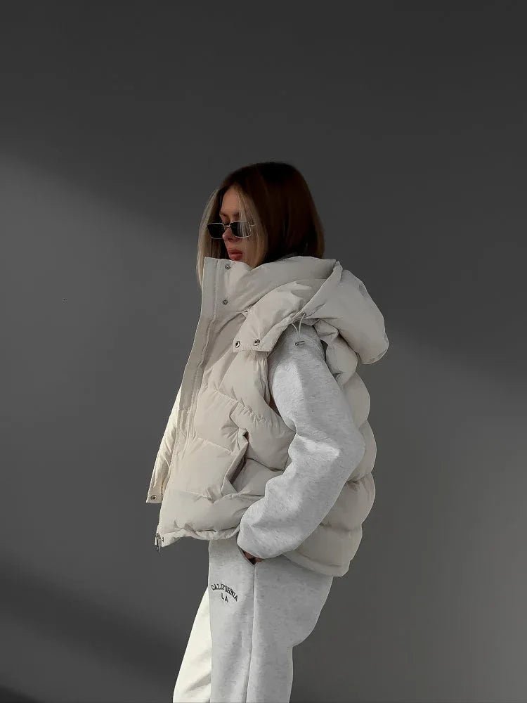 Three-quarter view of a woman wearing a cream puffer vest over a cream quarter-zip hoodie and cream joggers, with sunglasses and hands in pockets.