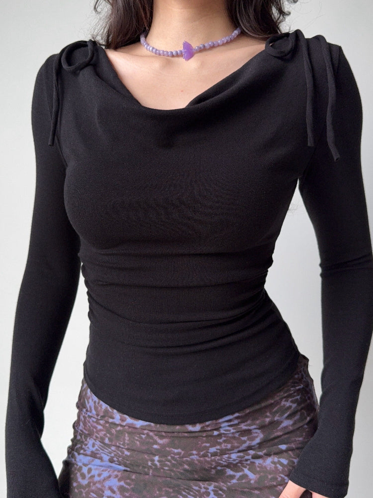 Full view of a black long-sleeve top with a draped neckline and ruched sides, styled with a necklace and patterned skirt.