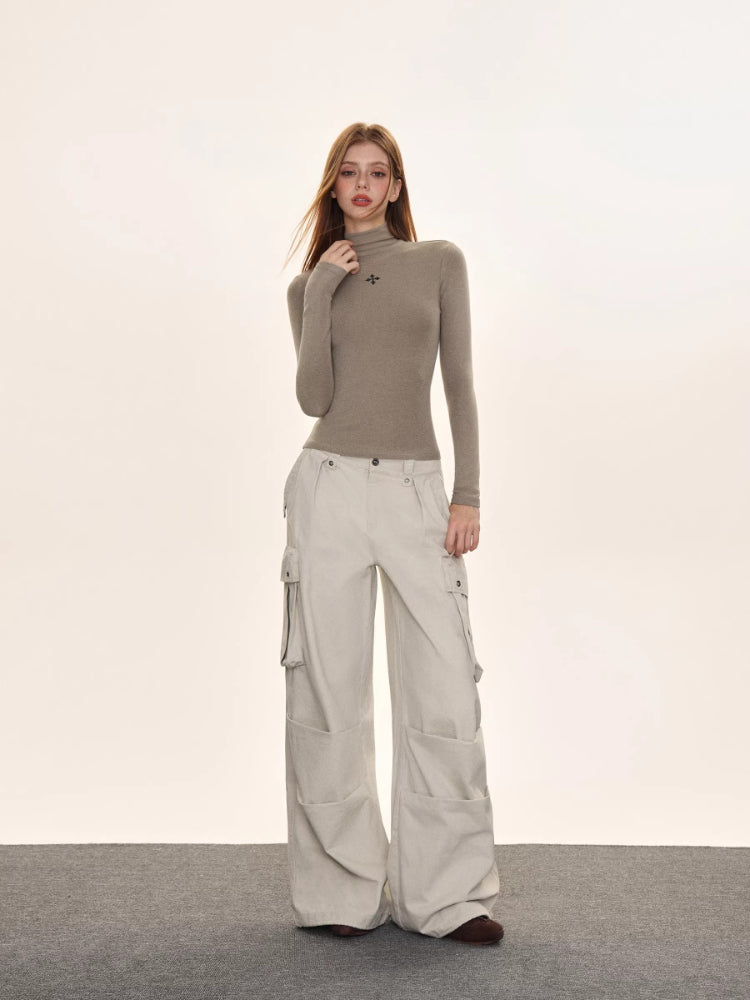 Full-body shot of a woman in a taupe turtleneck top with a black embroidered cross, and wide-leg light-colored cargo pants.