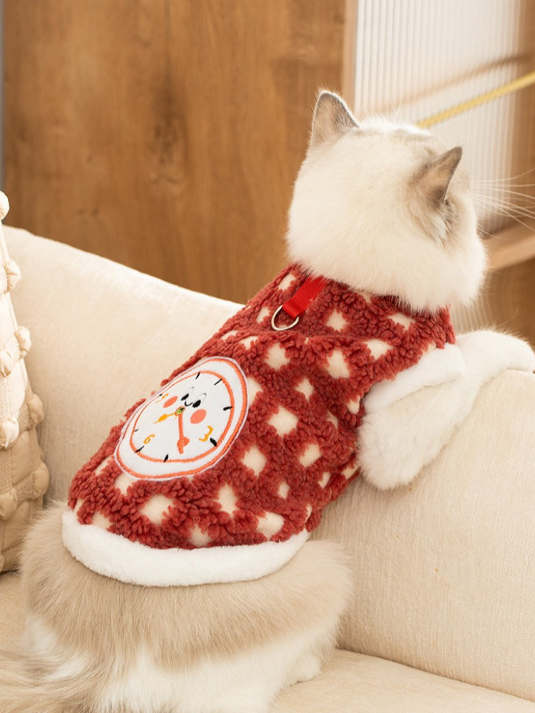 A fluffy white cat with blue eyes sits on a couch, facing away from the camera. The cat is wearing a red and white patterned fleece vest with a clock graphic on the back.