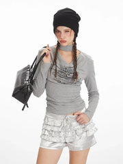 Model posing with a black handbag, wearing a grey long-sleeved top with a graphic print and silver ruffled shorts, black beanie, white socks, and black boots.
