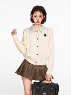 Woman wearing a cream-colored knit cardigan over a striped collared shirt, paired with a brown pleated skirt and a black handbag.