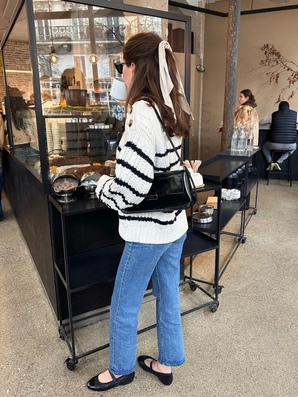 Woman standing at a cafe counter, wearing a striped knit sweater and blue jeans, with a handbag over her shoulder.