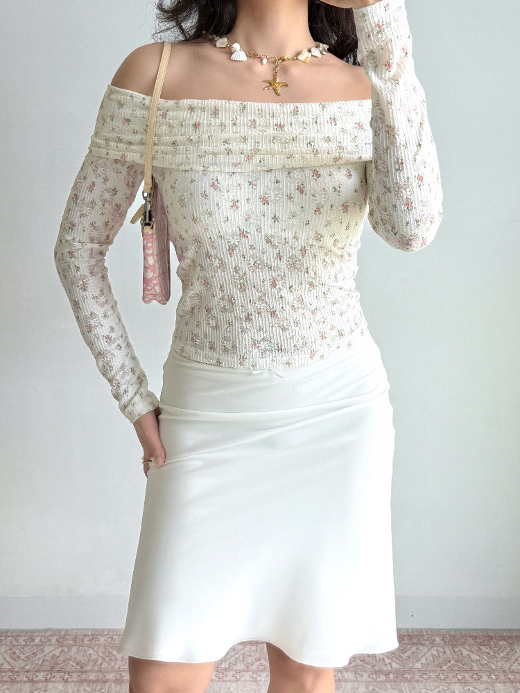 Woman wearing a white floral off-the-shoulder long-sleeved top, white slip skirt, seashell necklace, and a small pink handbag.