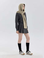 Full body shot of a model wearing a gray sweater and pleated skirt with a beige hooded scarf, black socks, and sneakers.