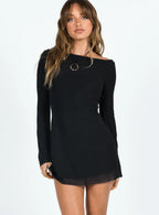 Close-up of a woman wearing a black long-sleeved mini dress with bell sleeves and a sheer hemline, with a gold pendant necklace.