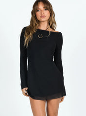 Close-up of a woman wearing a black long-sleeved mini dress with bell sleeves and a sheer hemline, with a gold pendant necklace.