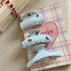 Three light blue and white striped hair claw clips with black bow prints arranged on a plaid surface next to a gift box.
