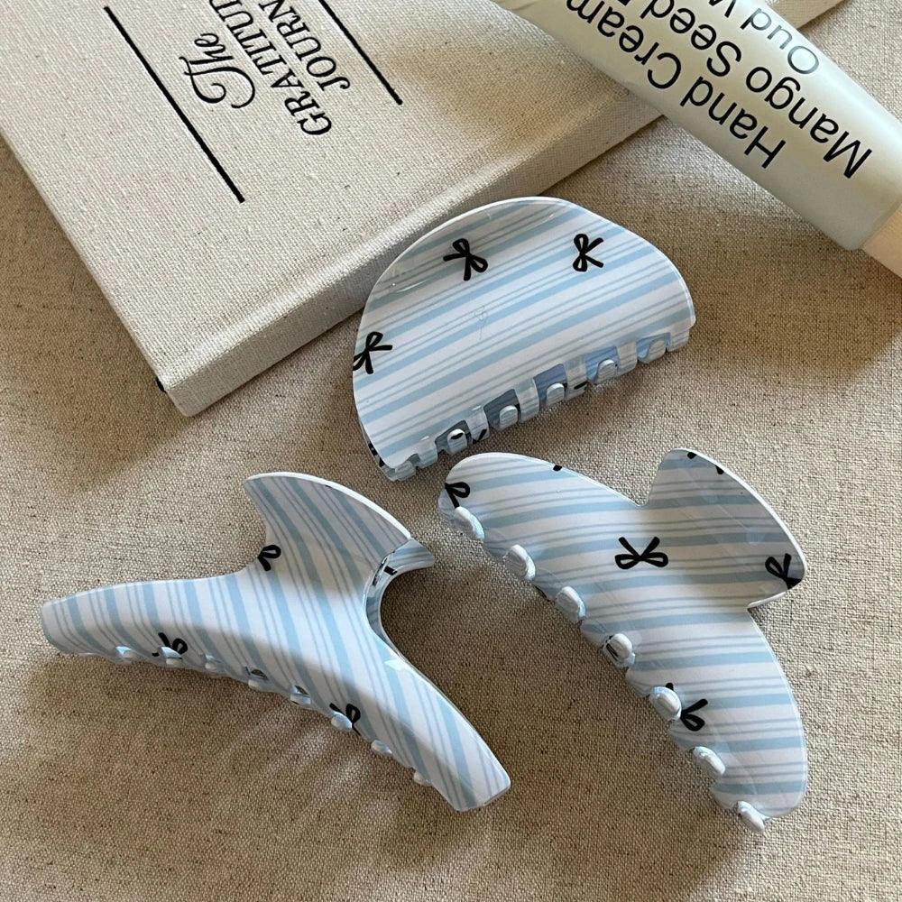 Three light blue and white striped hair claw clips with black bow prints arranged on a textured surface next to a book and a tube of hand cream.