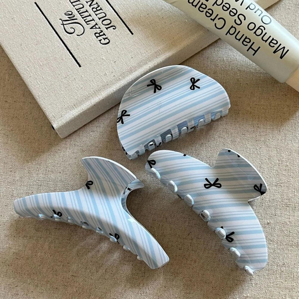 Three light blue and white striped hair claw clips with black bow prints arranged on a textured surface next to a book and a tube of hand cream. Secondary image