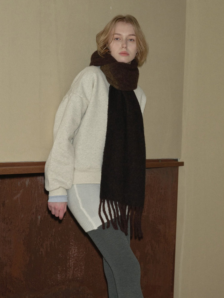A young person wearing a cream-colored crew neck sweatshirt and a dark brown fringed scarf, leaning to the side.