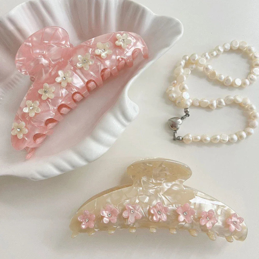 A pink marbled claw clip with floral and gemstone embellishments rests in a shell-shaped dish, next to a cream marbled claw clip with pink floral accents and a pearl necklace.
