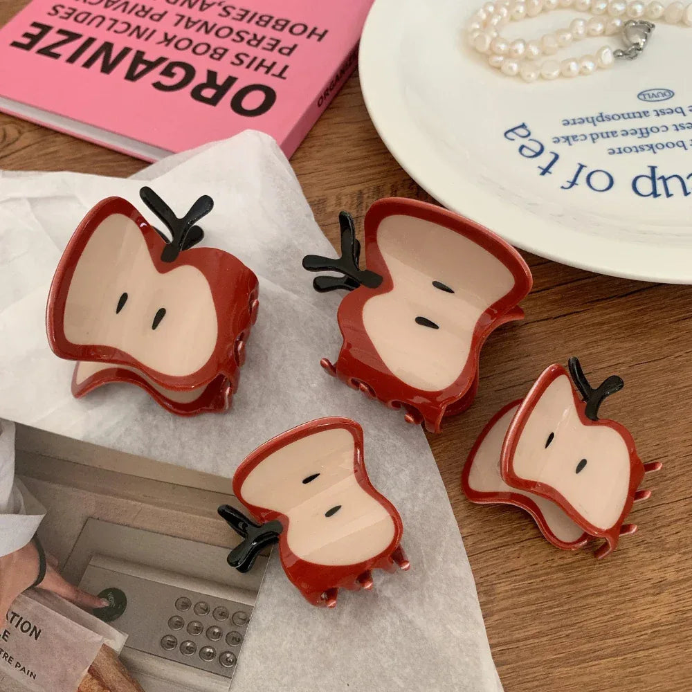 A close-up of four apple slice hair claw clips scattered on a white textured surface, with a pink book and a plate in the background.