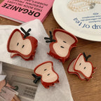 A close-up of four apple slice hair claw clips scattered on a white textured surface, with a pink book and a plate in the background.