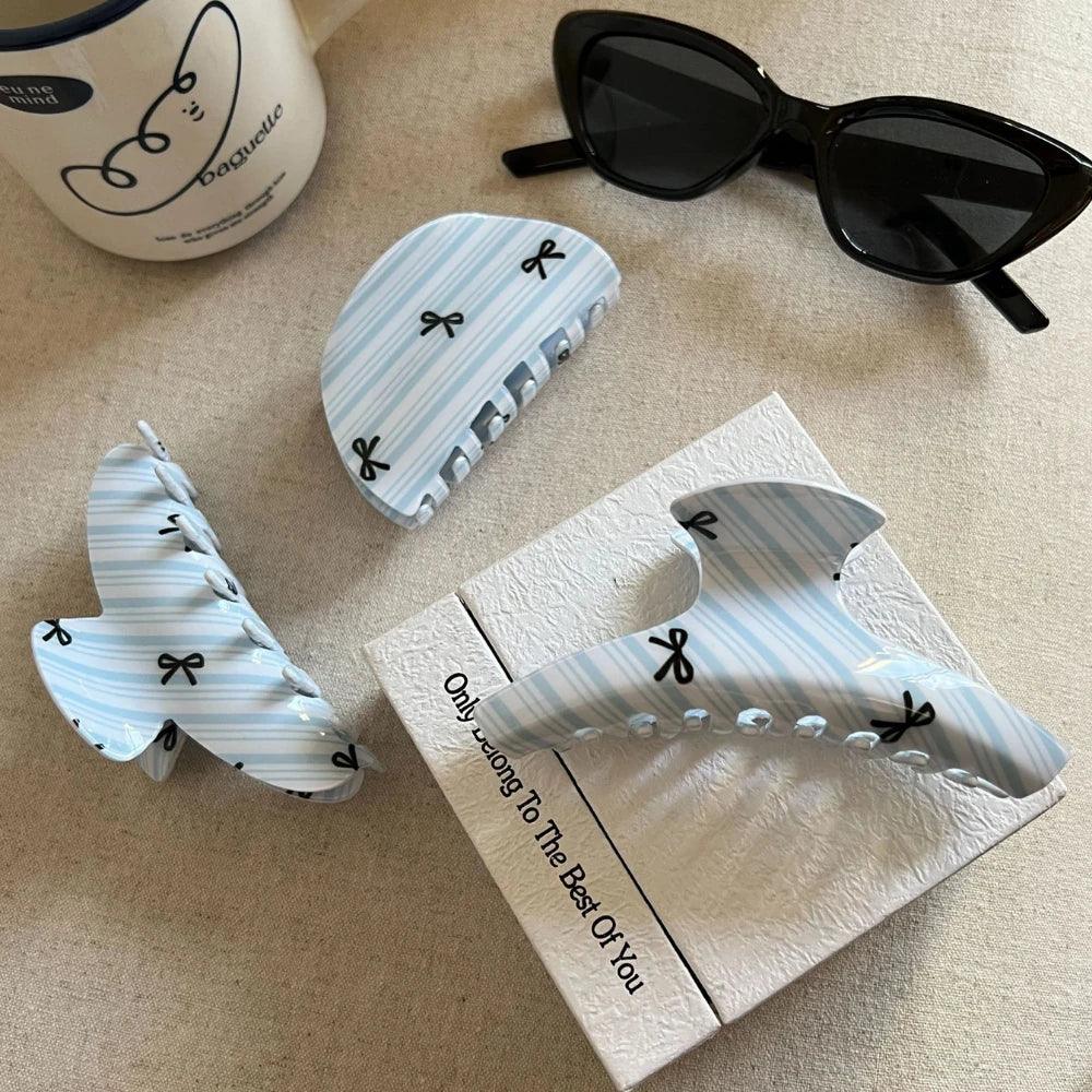 Three light blue and white striped hair claw clips with black bow prints arranged on a box, next to a mug and sunglasses.