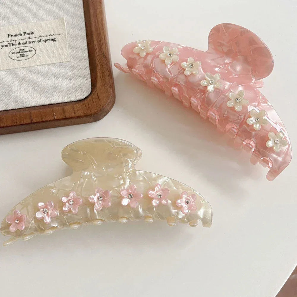 Two marbled claw clips, one pink and one cream, both embellished with floral details and gemstones, arranged on a white surface next to a framed print.
