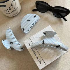 Three light blue and white striped hair claw clips with black bow prints arranged on a box, next to a mug and sunglasses.