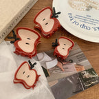 Four apple slice hair claw clips of varying sizes arranged on a wooden surface, with books and a plate in the background.
