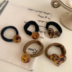 Group of leopard print hair ties with round and heart charms, placed on a fabric with text.