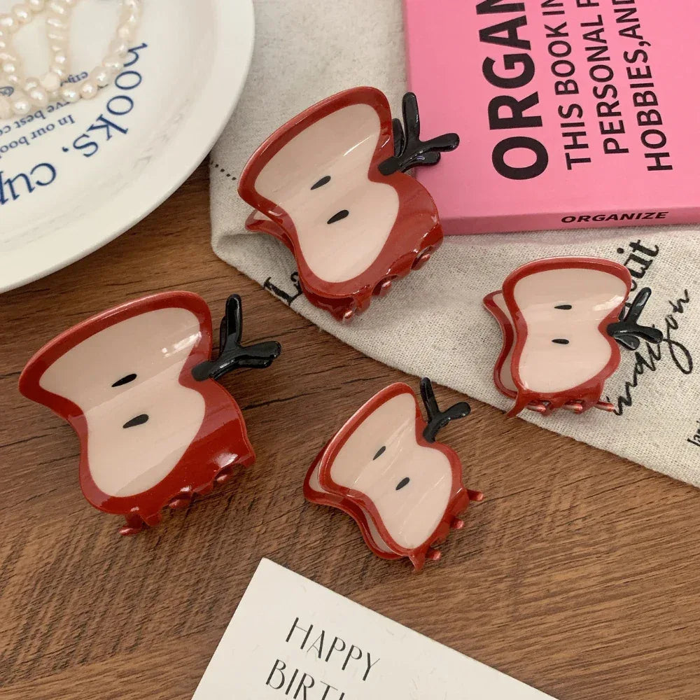A collection of four apple slice hair claw clips displayed on a wooden surface, with various decorative items including a book and a plate.