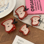 A collection of four apple slice hair claw clips displayed on a wooden surface, with various decorative items including a book and a plate.